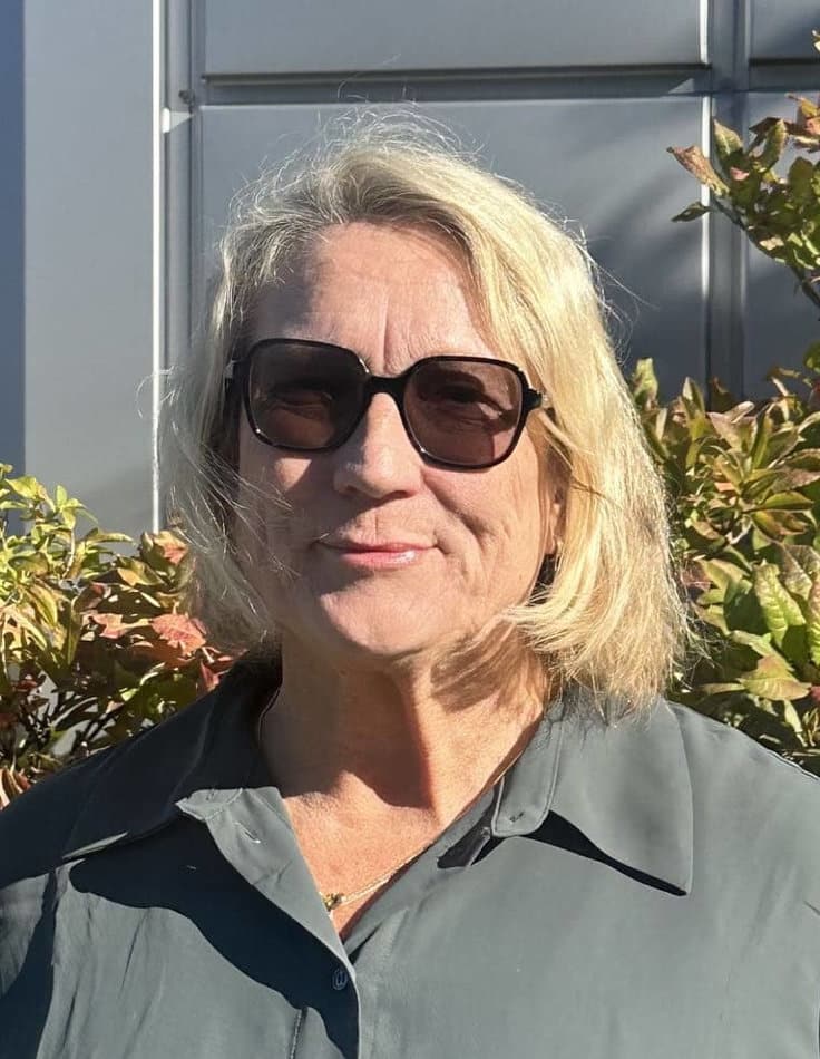 Kvinna med kort blont hår och solglasögon står utanför, iklädd en mörkgrön skjorta. Solbelyst bladverk och en byggnad med metallisk fasad finns i bakgrunden.